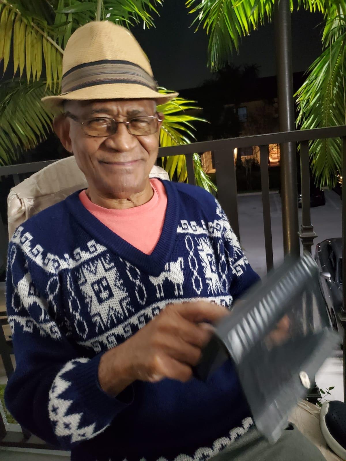 Elderly man wearing a hat and patterned sweater, sitting outdoors near palm trees at night, holding an envelope or booklet.