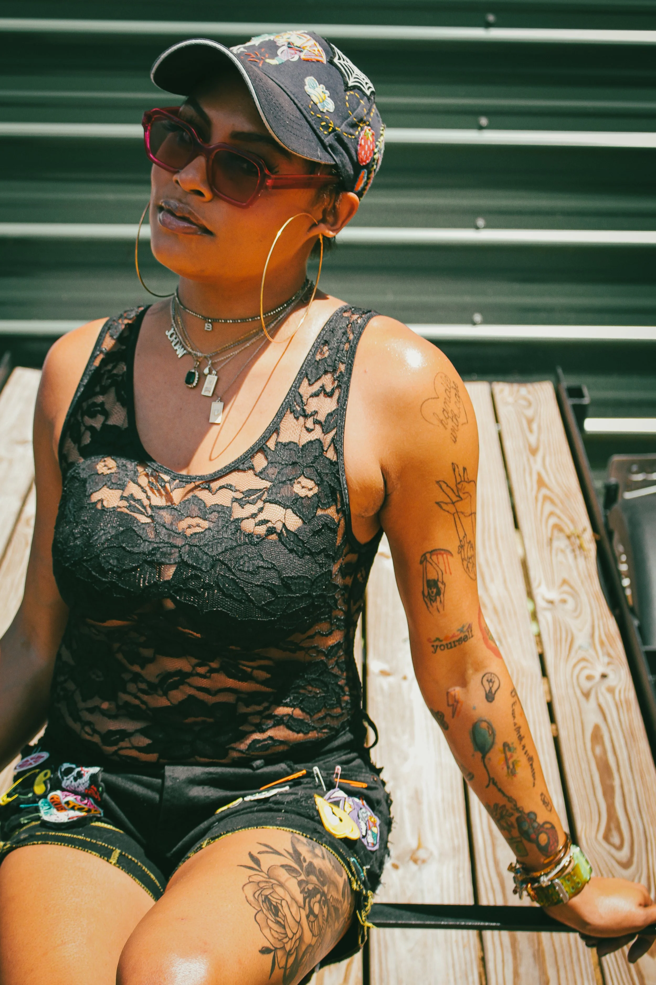 A woman sitting on a wooden bench outdoors, wearing sunglasses, a black lace top, and colorful shorts, with tattoos on her arms and thigh, and a black cap with embroidery.