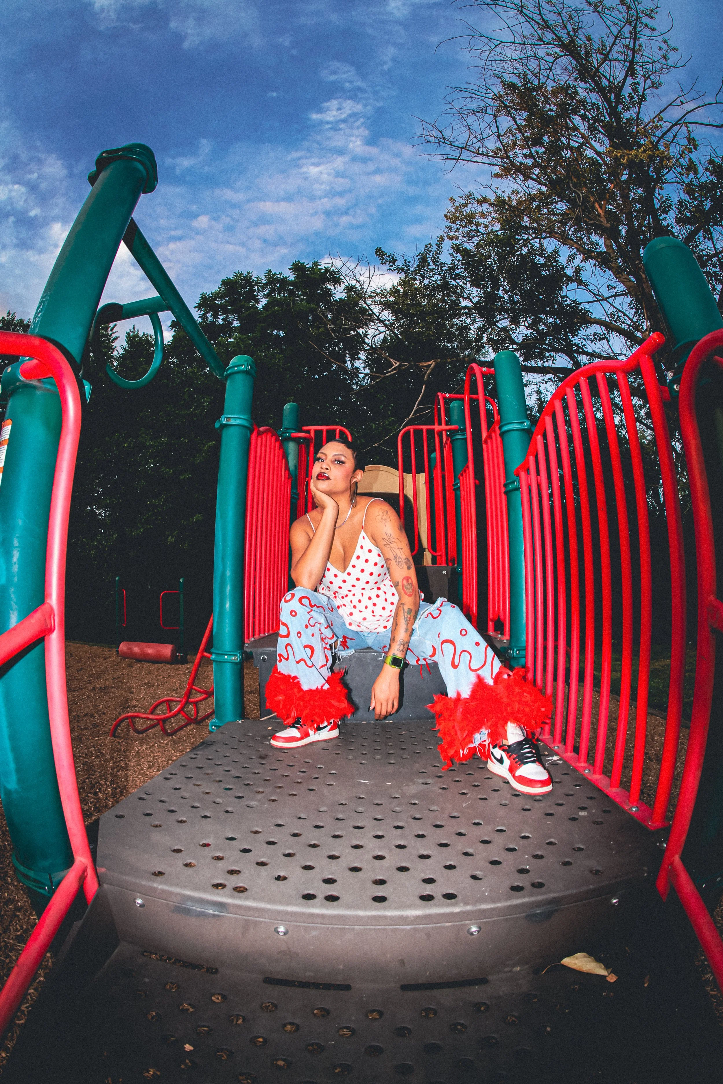 A woman with tattoos and dark makeup sitting on playground equipment at dusk, wearing a polka dot top, light blue pants with red squiggles, red feathered shoes, and earrings, surrounded by trees and blue sky.