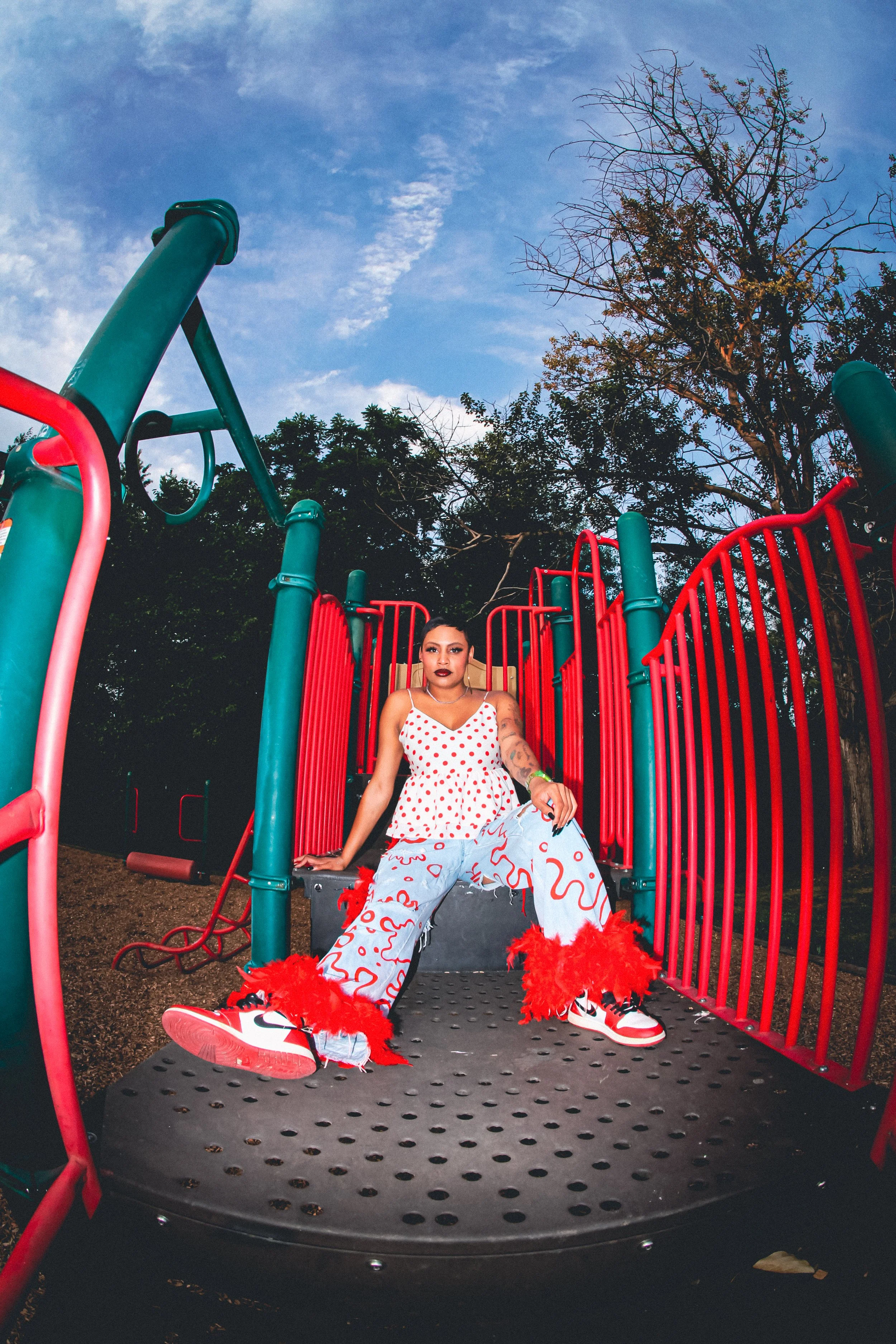 Person sitting on a playground structure wearing a white top with red polka dots, patterned pants, red furry shoes, and red lipstick, with trees and a blue sky in the background.