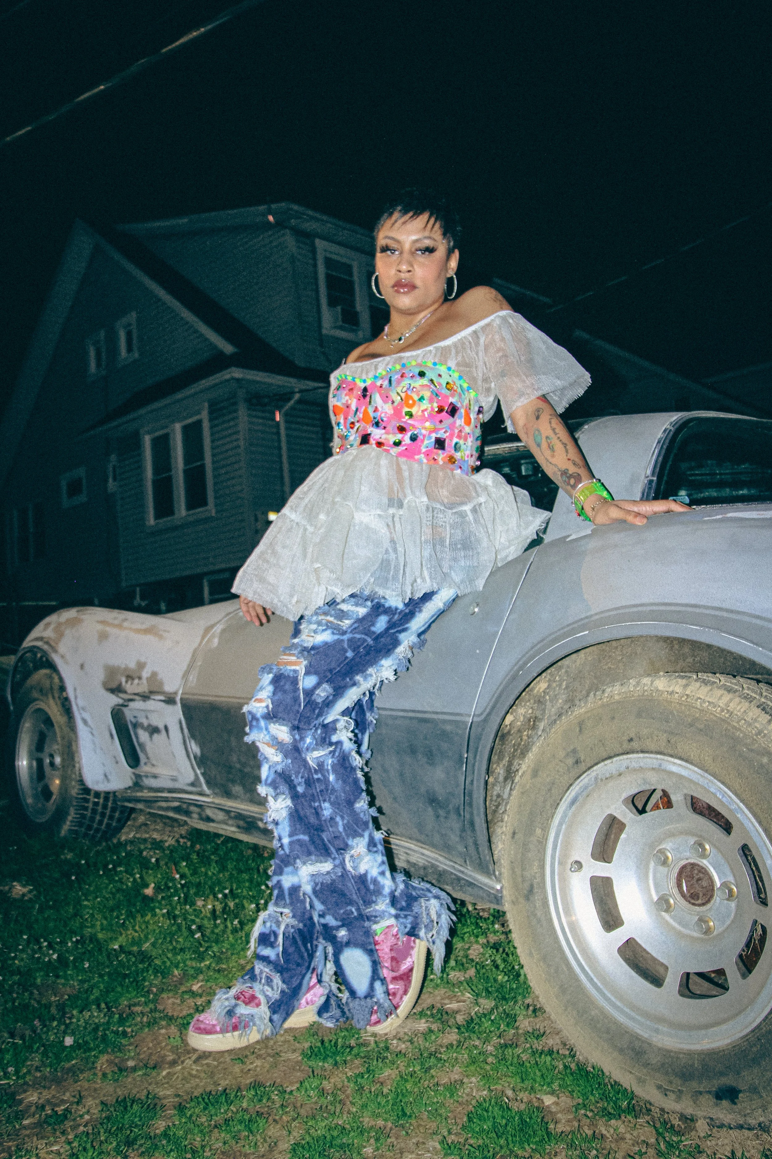 A woman with short dark hair and tattoos on her arm, wearing a colorful off-shoulder top and ripped jeans, sitting on a vintage car in a residential neighborhood at night.