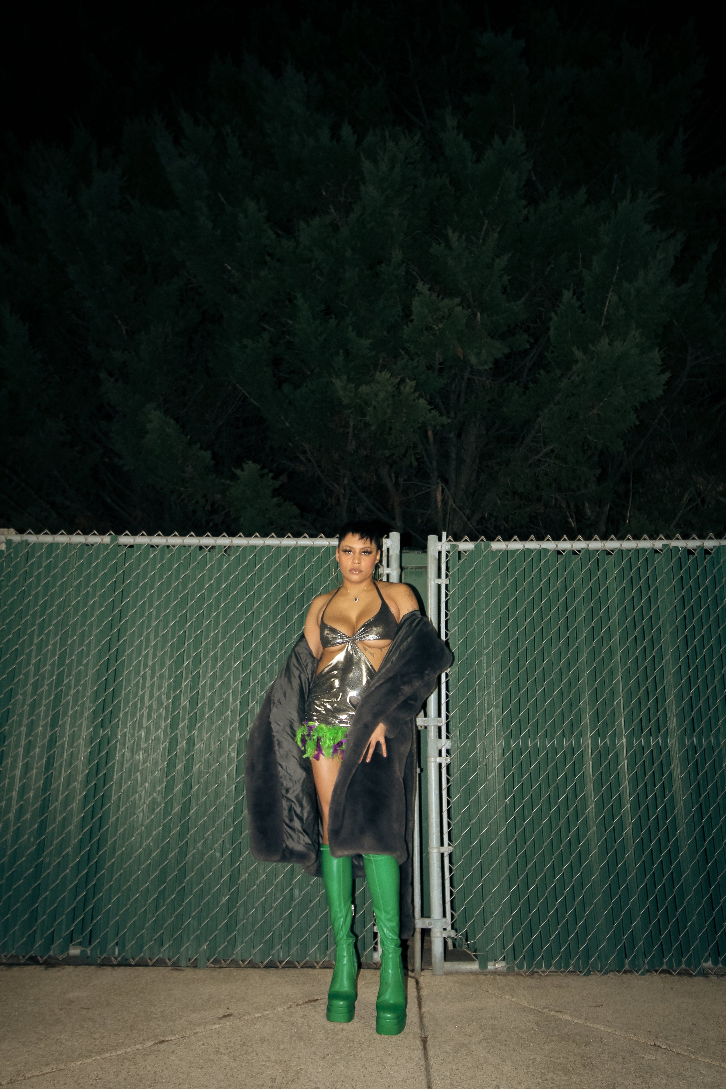 A woman standing in front of a green chain-link fence at night, wearing a metallic silver dress, green thigh-high boots, and a black coat draped off her shoulders.