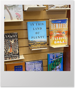 Display of various books on shelves in a bookstore, featuring titles like 'In This Land of Plenty’.