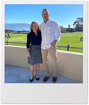 A man and woman posing together outdoors in front of a scenic landscape with grass, mountains, and a clear blue sky.