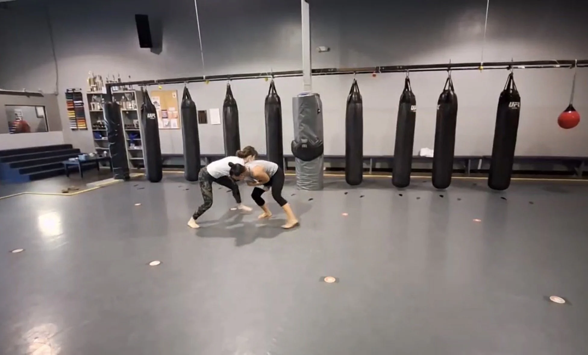 Two women practicing Brazilian Jiu-Jitsu in an empty martial arts gym with punching bags hanging on the wall.