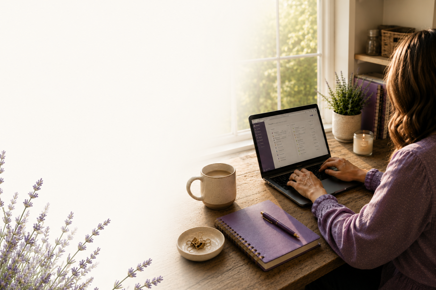 Woman working on a laptop at a wooden desk near a window, with a purple notebook, pen, coffee mug, purple flowers, a small bowl, and a candle.