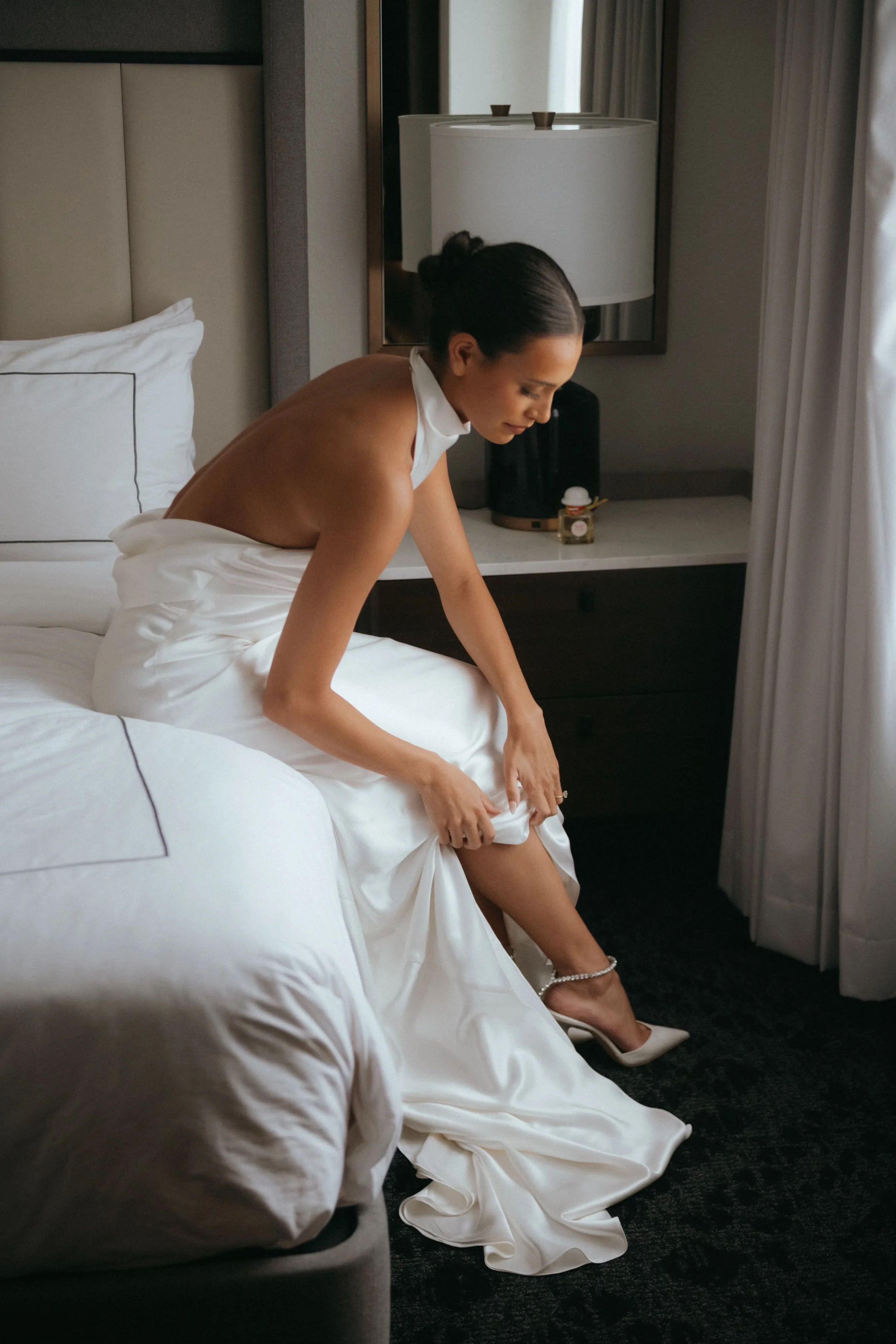 Woman in a bridal gown putting on high-heeled shoes in a hotel room.