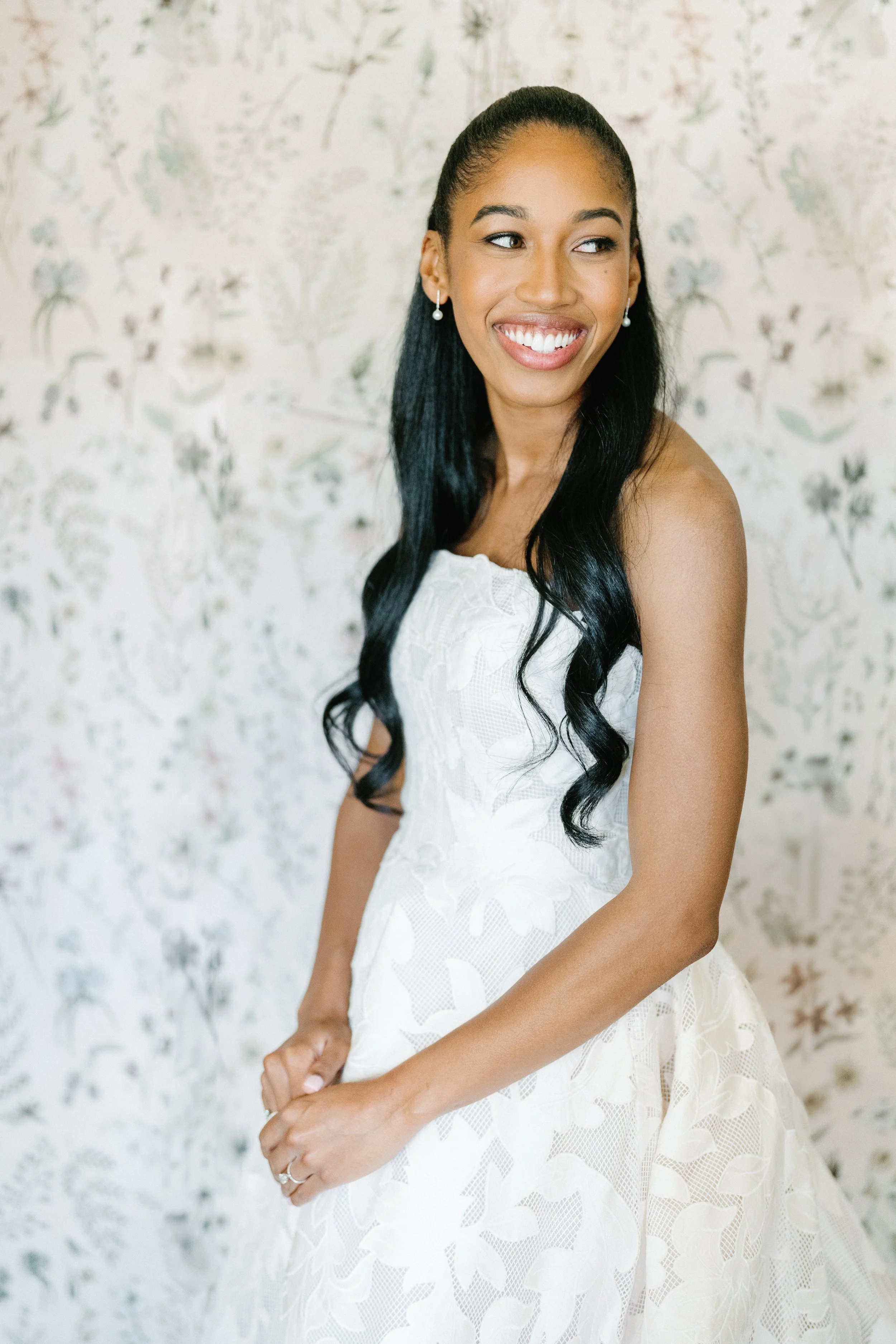 A woman in a white dress smiling, with long black hair, in front of a floral wallpaper background.