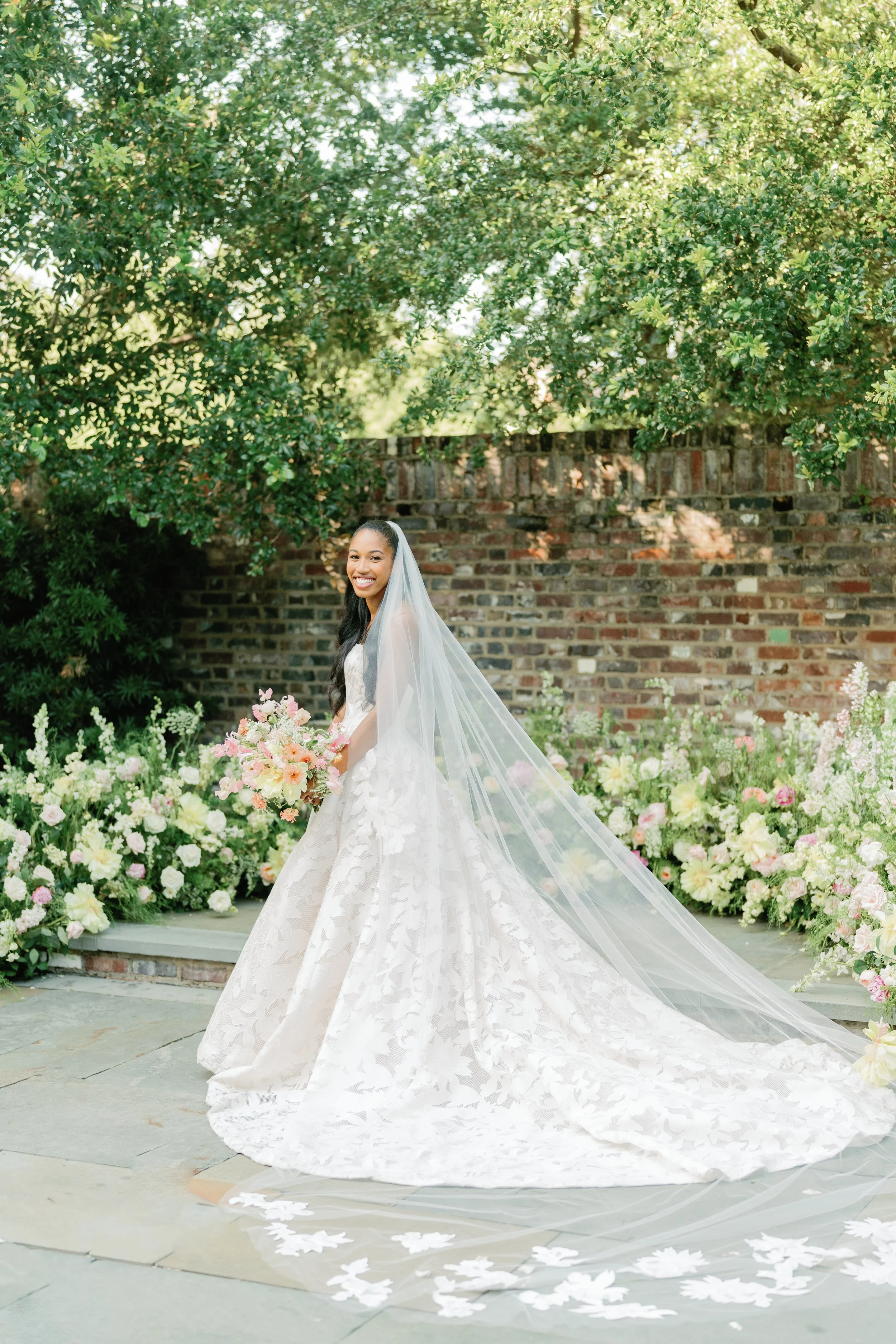 A bride in a white wedding gown walking outdoors, holding a bouquet of pink and peach flowers, surrounded by lush greenery and a brick wall, with a long veil trailing behind her.