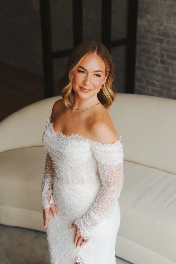 A woman in an off-shoulder white lace wedding dress, standing in front of a white sofa with a brick wall background.