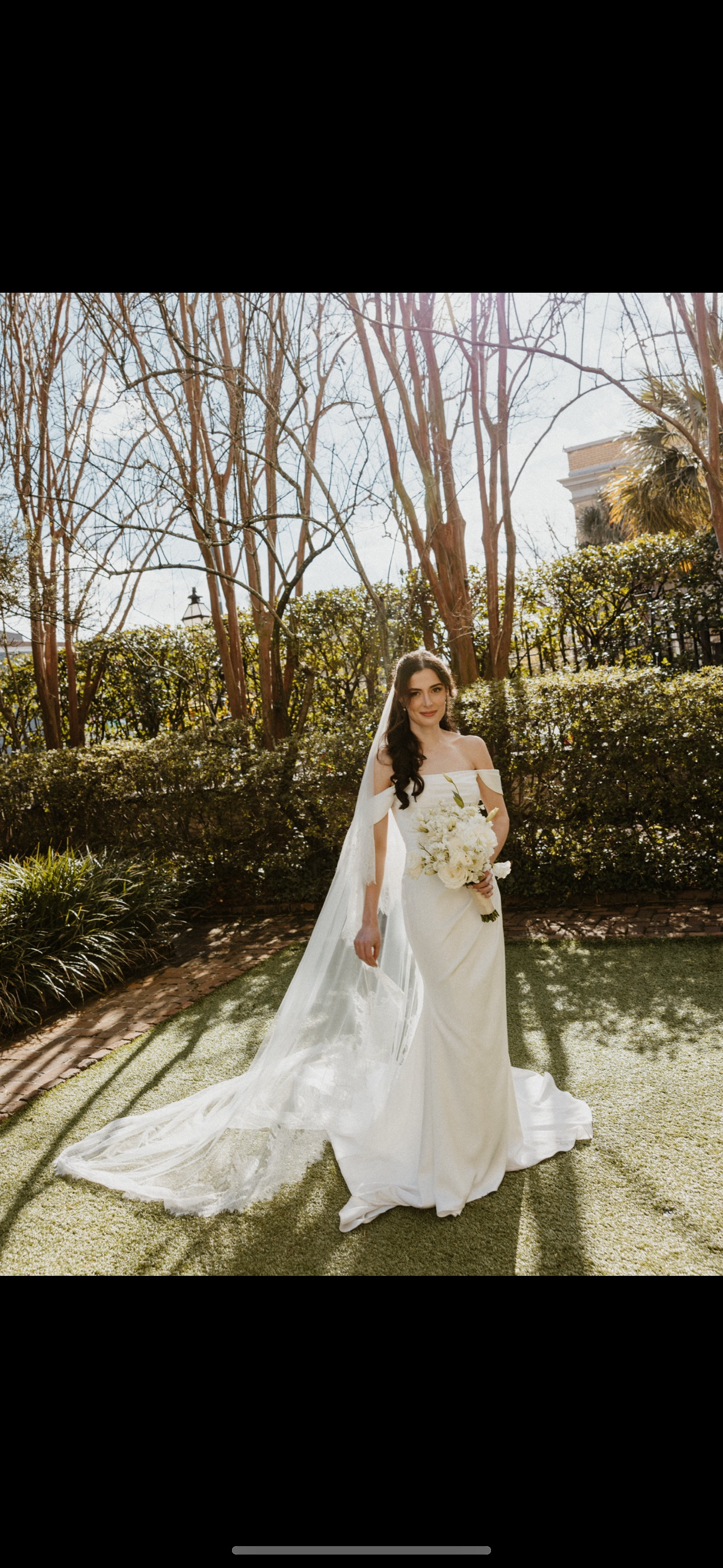 A bride in a white wedding gown standing outdoors on a lawn, holding a bouquet of flowers, with sunlight and trees in the background.