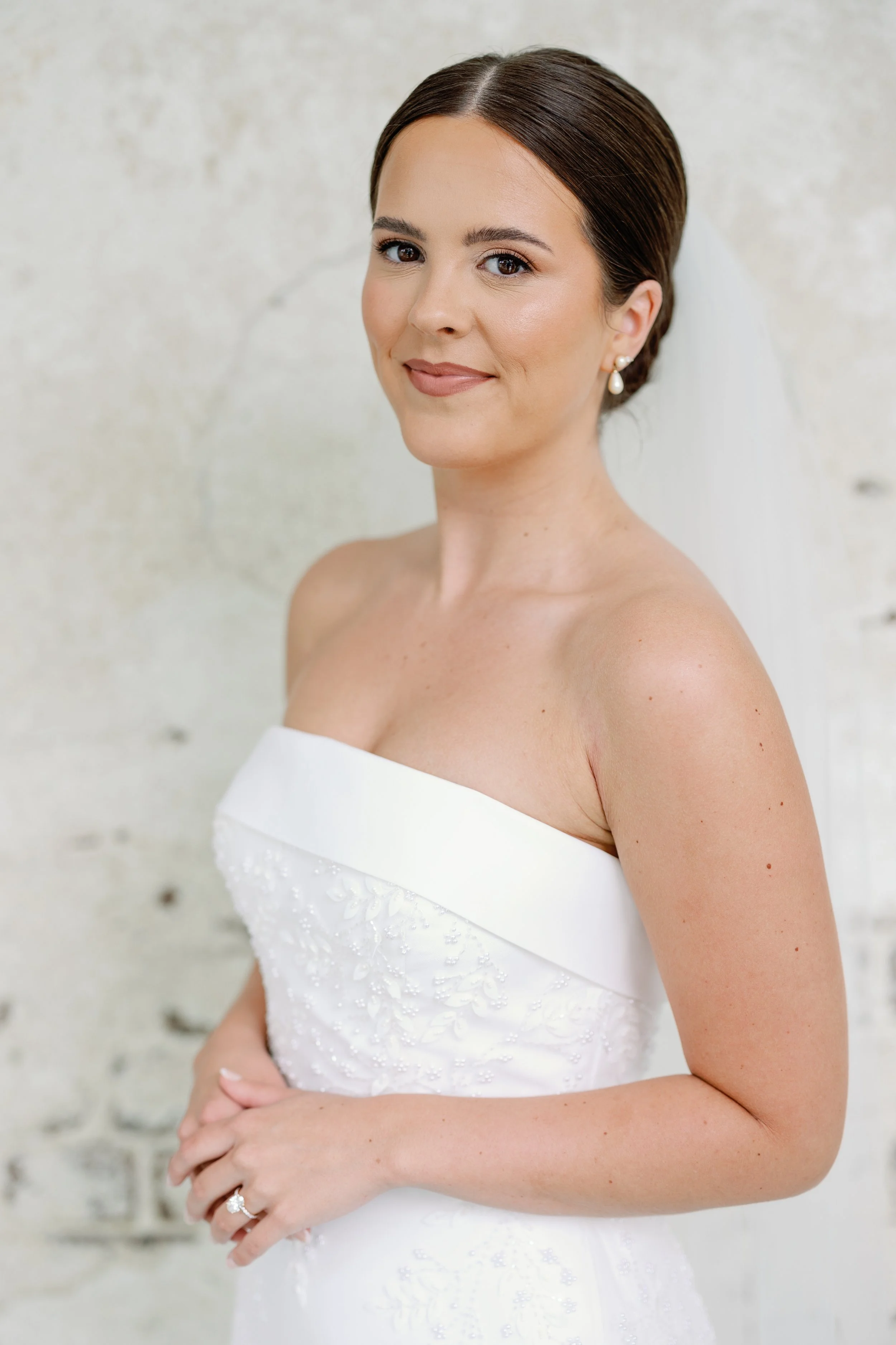 A bride with brown hair in a white strapless wedding dress standing against a light-colored textured wall.