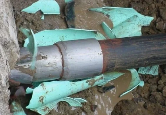 Broken green plastic pieces surrounding a gray and black metal pipe buried in soil.