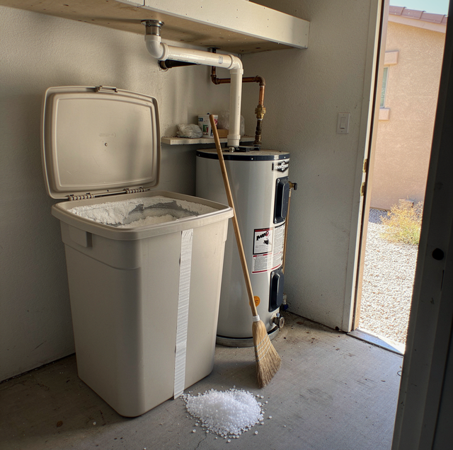 Trash can and water heater in a utility space with broom and salt pile on the floor.