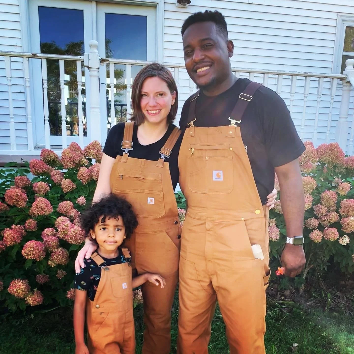 A family of three standing outdoors in front of flowers and a white house, all wearing tan Carhartt overalls.