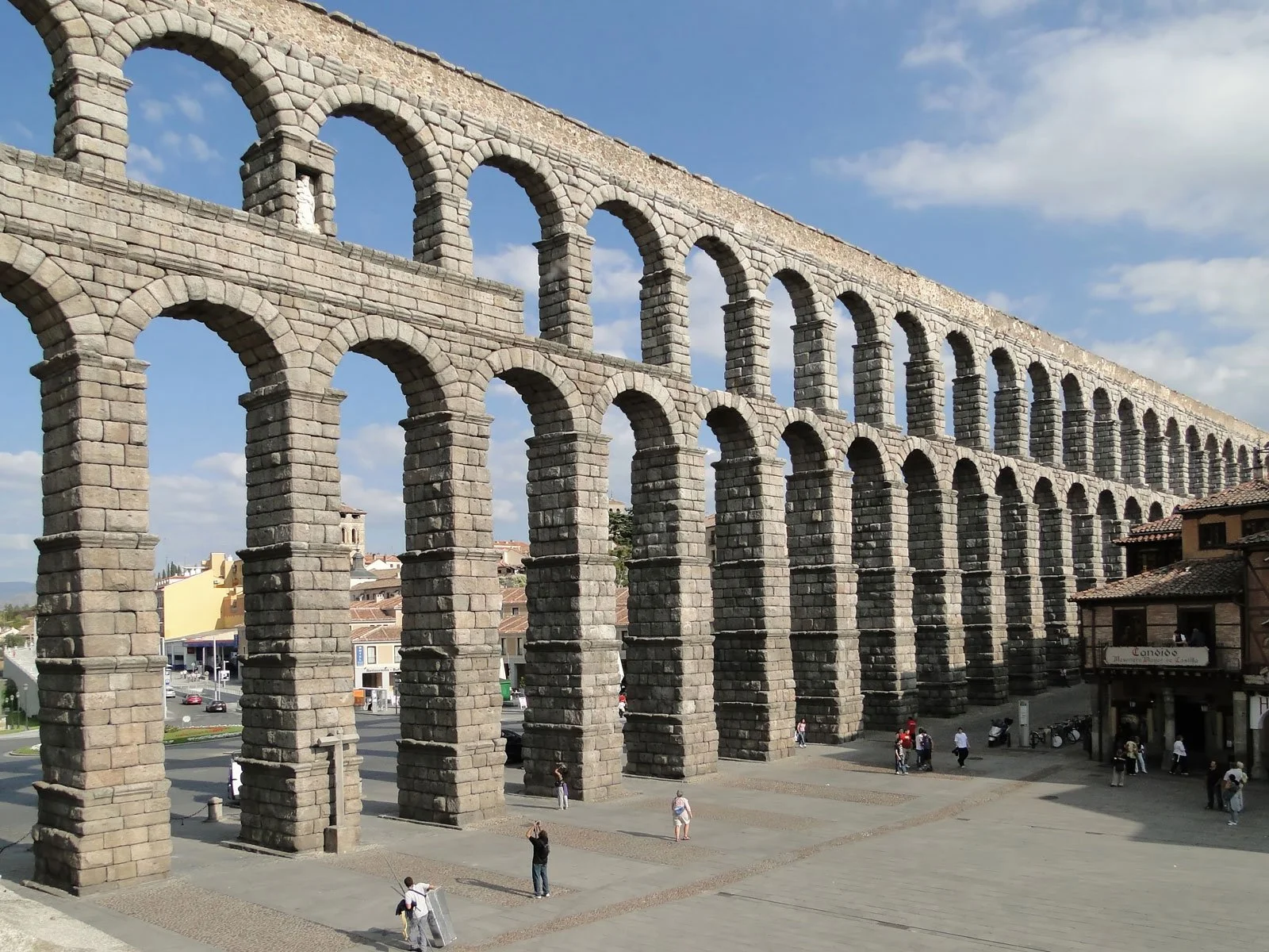 Wide urban view of the Aqueduct of Segovia with multiple tiers of arches crossing a city square.