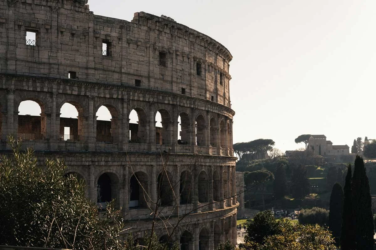 The Colosseum in Rome seen from the side, with ancient stone arches, trees, and soft light over the Roman ruins.