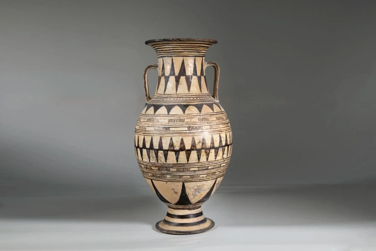 Tall Proto-Geometric amphora with twin handles, black bands and repeating triangular and linear motifs on pale clay.