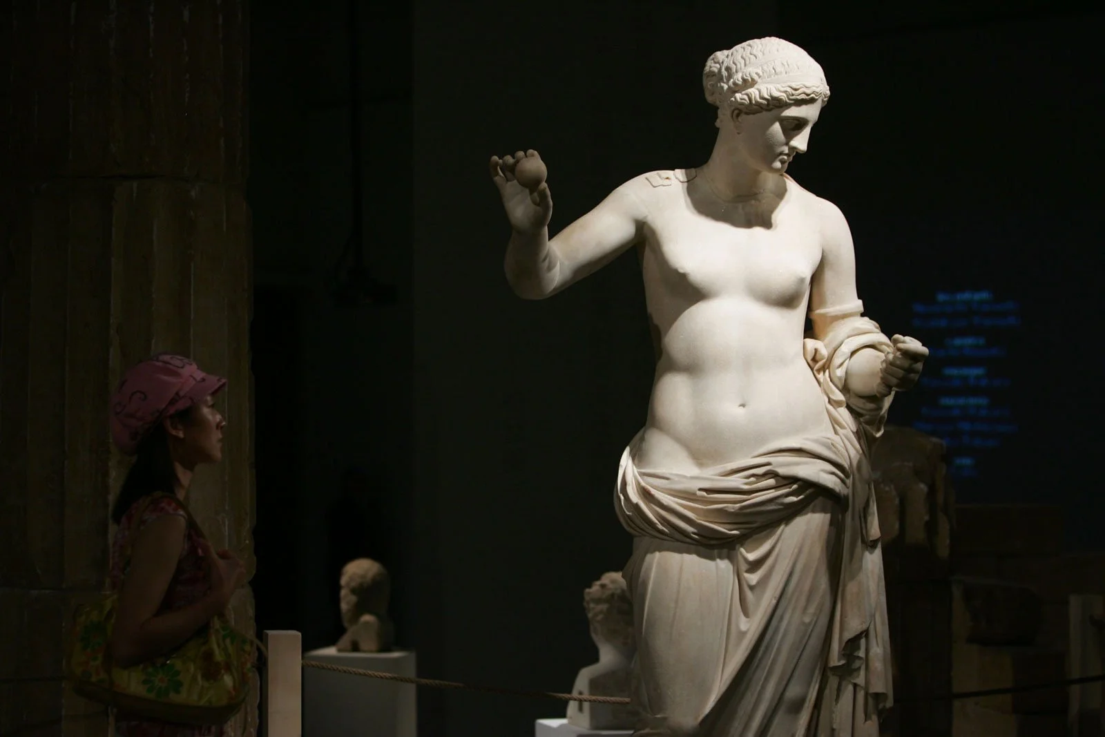 Marble Venus-type statue in a dark museum gallery with a visitor standing nearby.