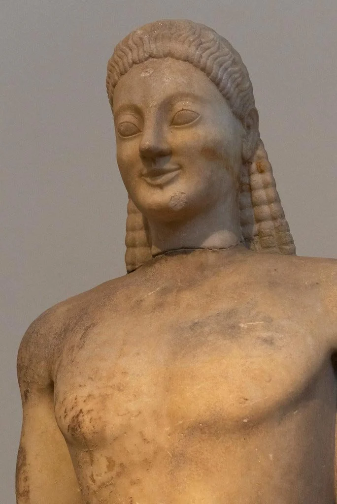 Marble kouros torso in close-up, with archaic smile, almond eyes and beaded hair, chest and shoulders softly modeled.