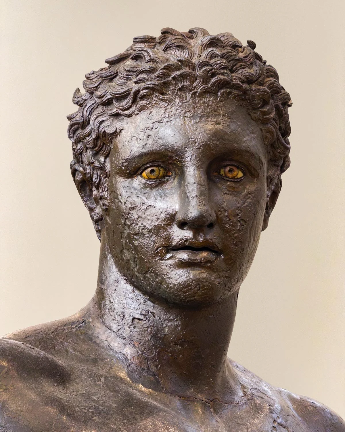 Close-up of a bronze youth’s head with vivid inlaid eyes and textured hair.