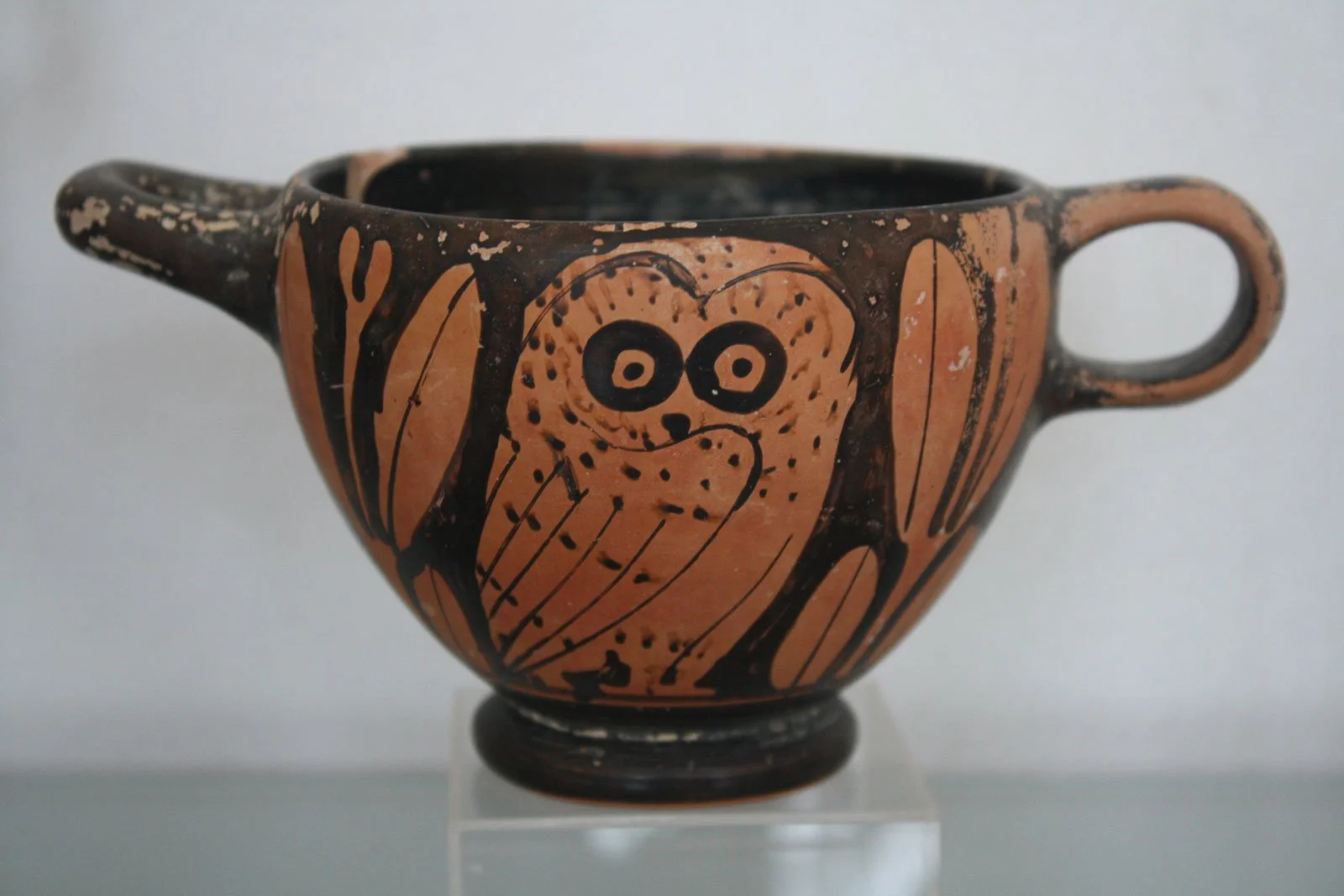 Small Greek drinking cup painted with a wide-eyed owl framed by stylized leaves, a playful, everyday nod to Athena, goddess of wisdom and of Athens.