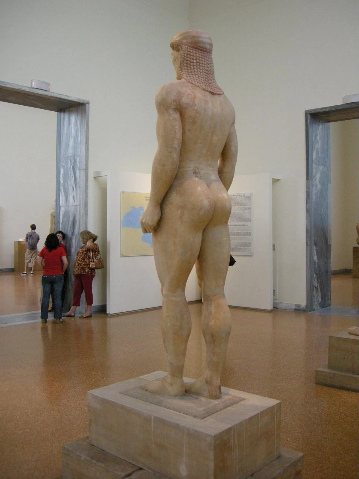 Back view of the Anavysos Kouros in museum gallery, showing powerful legs, rounded muscles and heavy braids hanging down the spine.