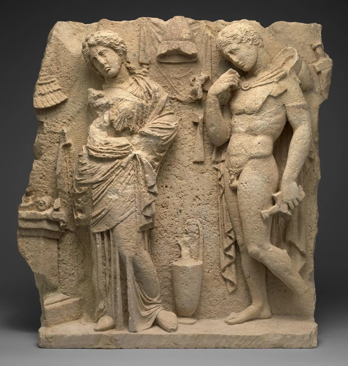 Stone relief showing a draped woman and a nude youth in contrapposto with carved objects around them.