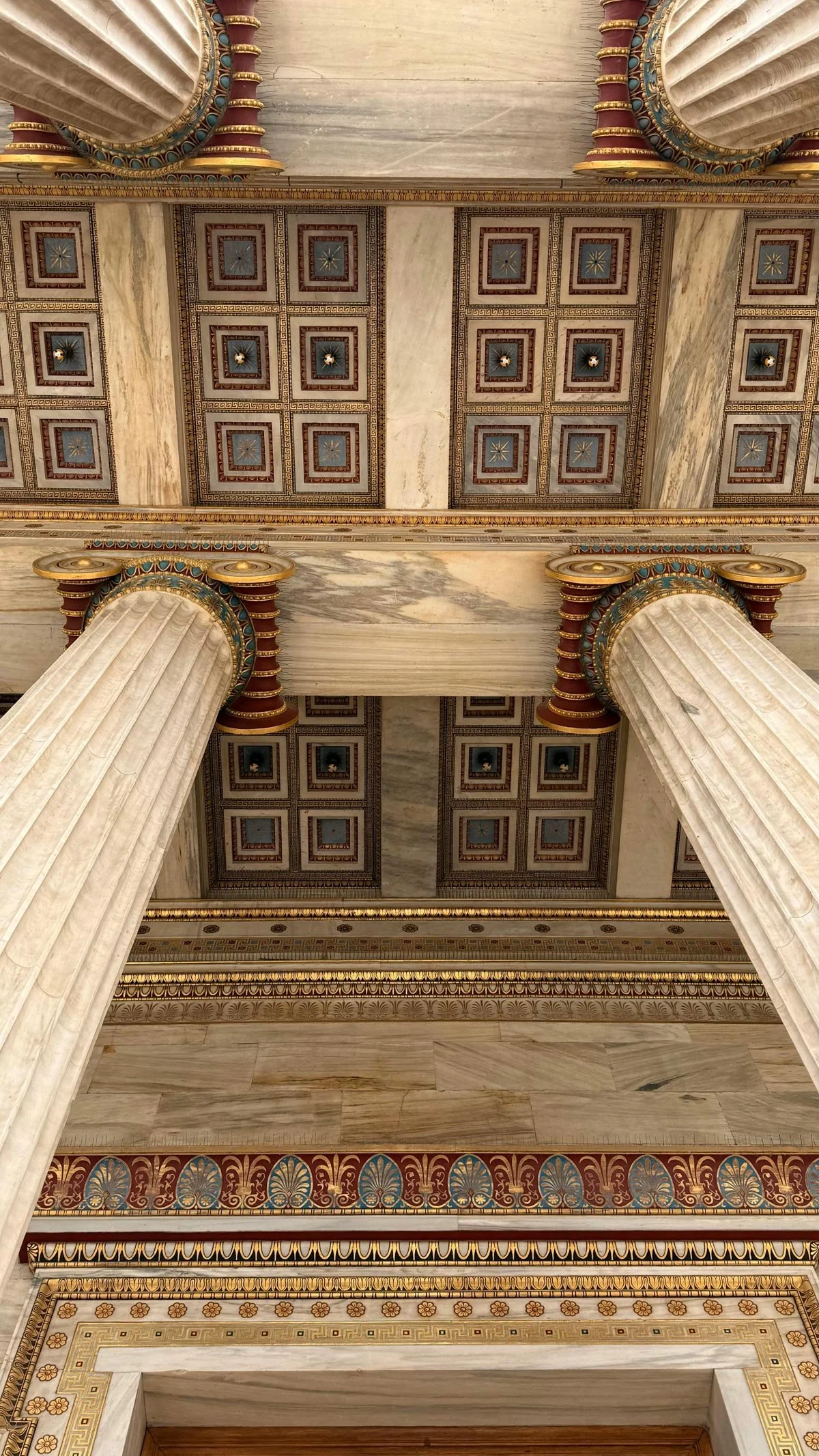 Upward view of a richly painted coffered ceiling with Ionic columns, starry panels and gold ornament recalling ancient polychromy.
