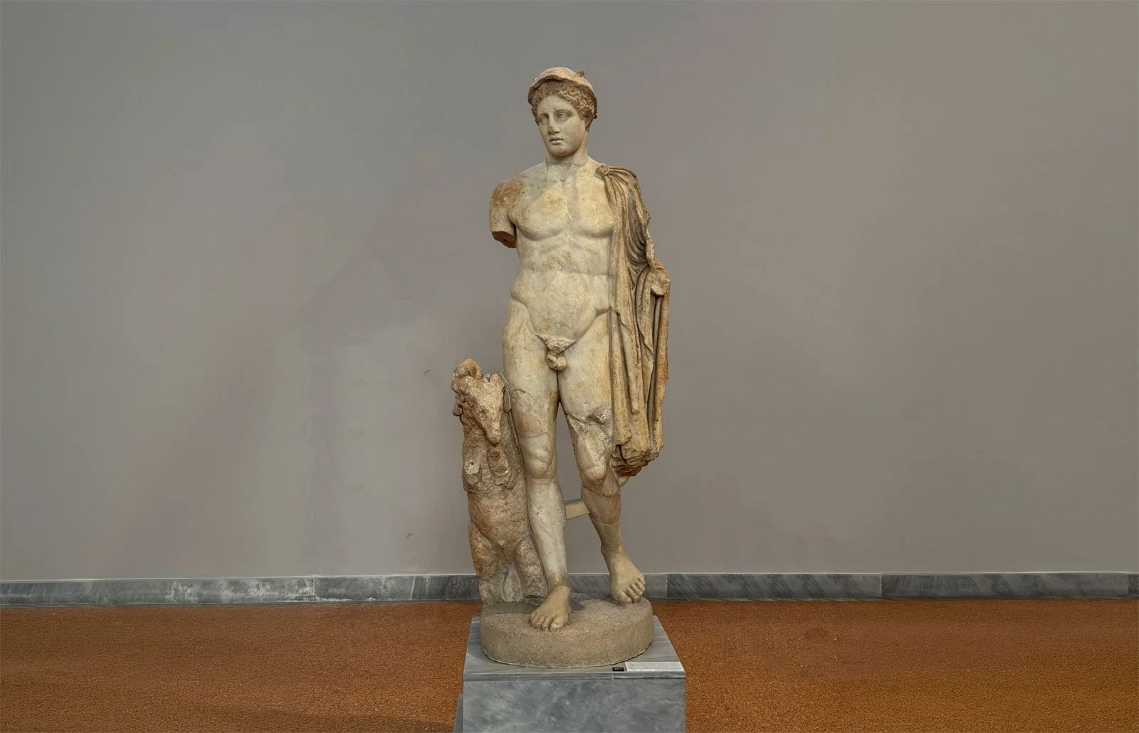 Full-length view of a marble nude male statue with missing arms displayed on a museum pedestal.