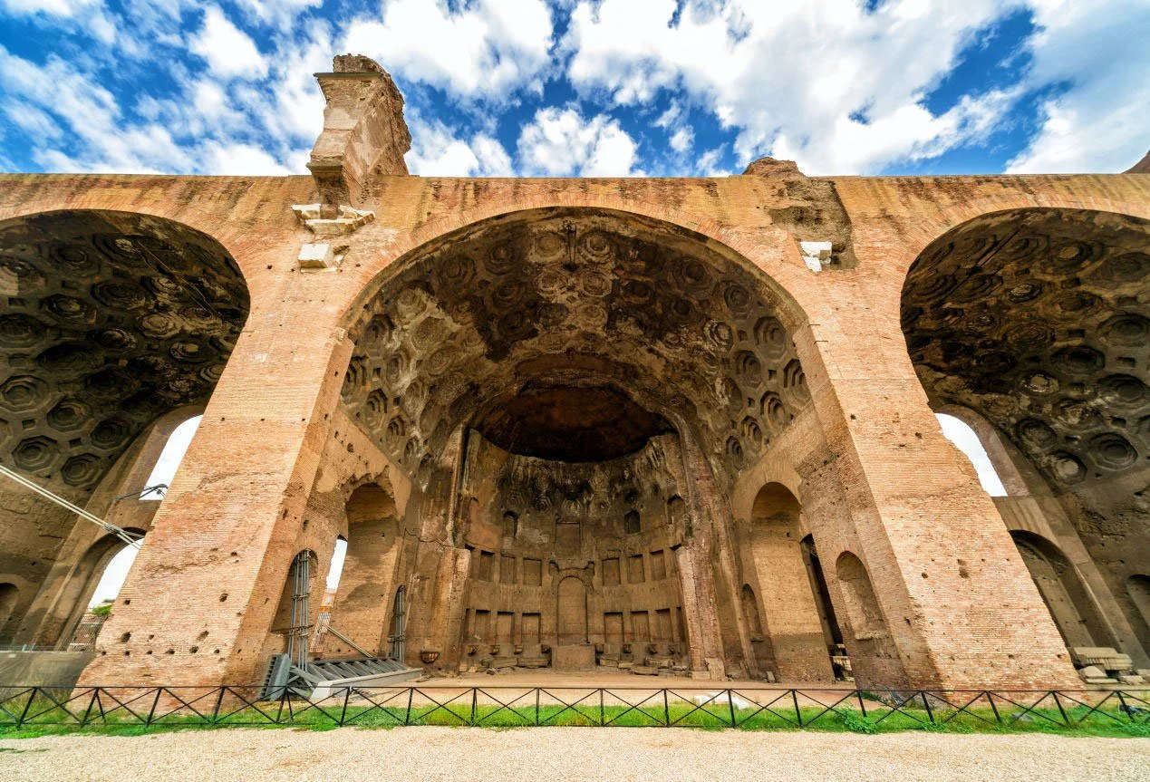 Ancient Roman basilica ruins with massive brick arches, vaulted ceilings, and a bright cloudy sky above.