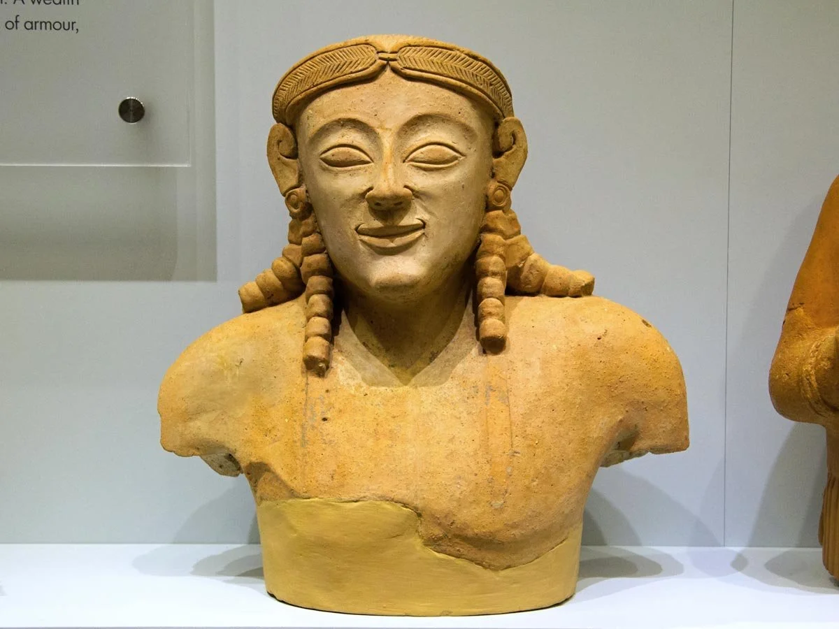 Terracotta kouros torso from Praisos, with archaic smile, stylised braids and simplified chest preserved as a compact bust.
