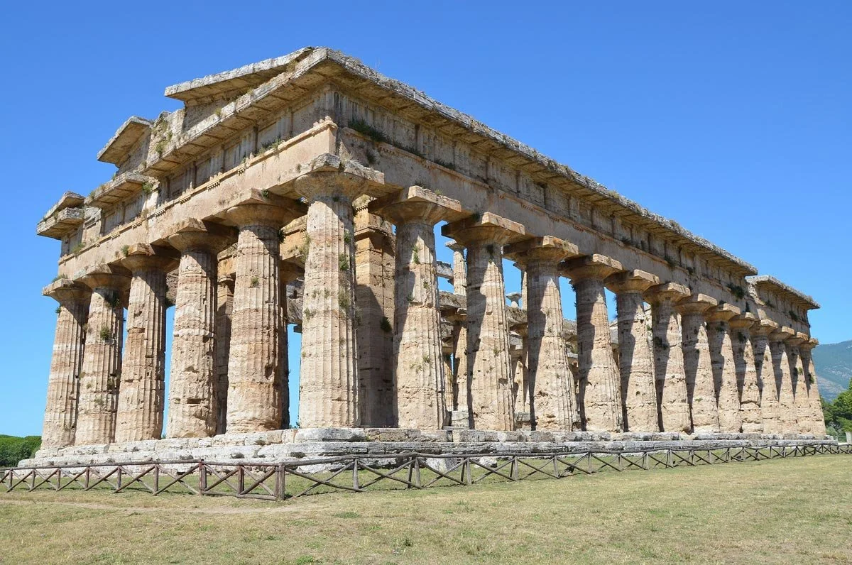 Side view of a well-preserved Doric temple at Paestum, its heavy fluted columns standing under a bright blue sky.