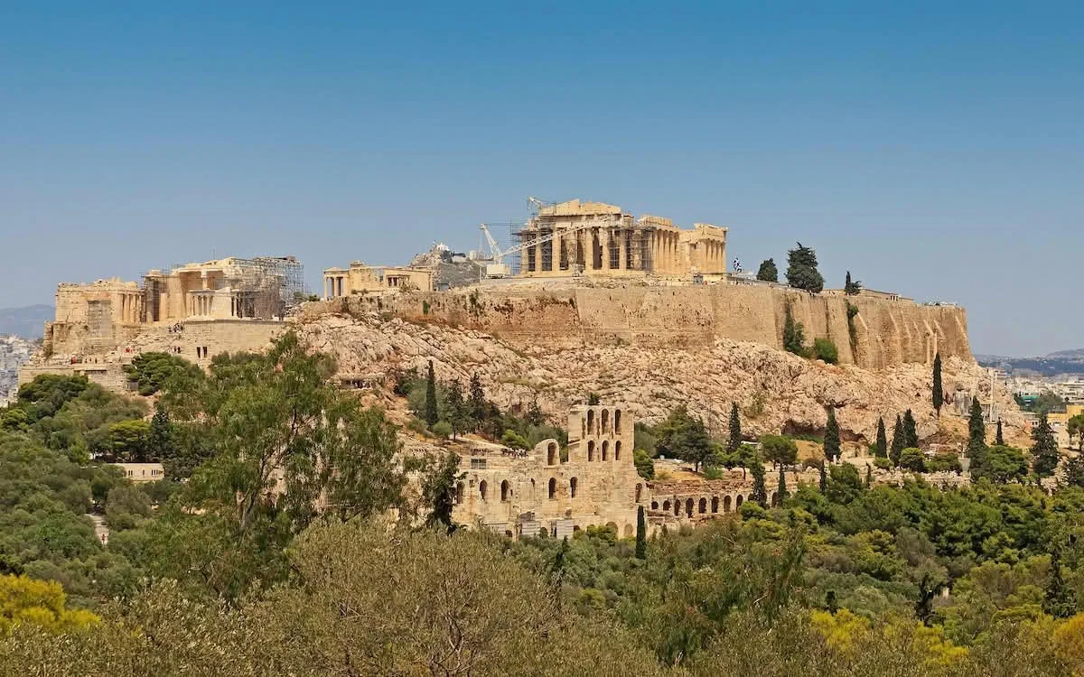 Distant panoramic view of the Acropolis of Athens, with the Parthenon crowning a rocky hill above green trees and city.