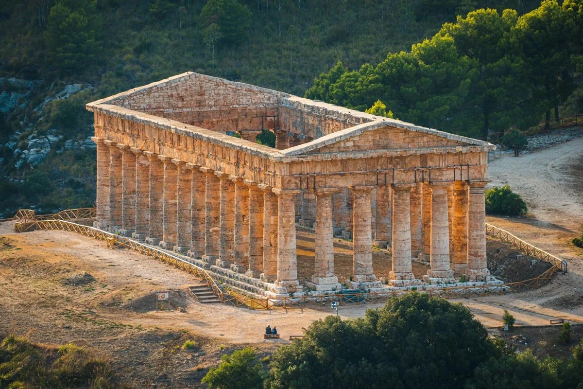 Aerial view of the Doric temple of Segesta on a hilltop, surrounded by paths, trees and warm Mediterranean light.