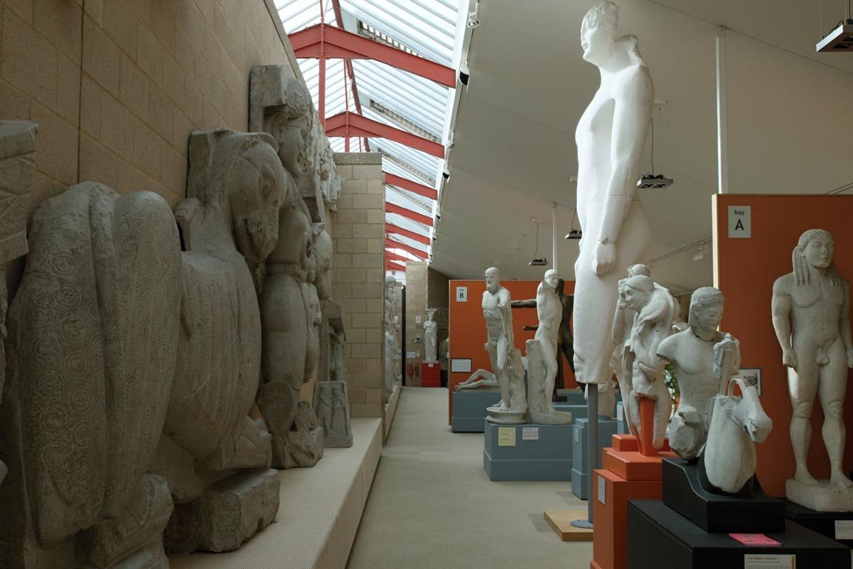 Gallery of plaster casts of archaic Greek sculptures, lined along a high-lit museum corridor with skylights above.