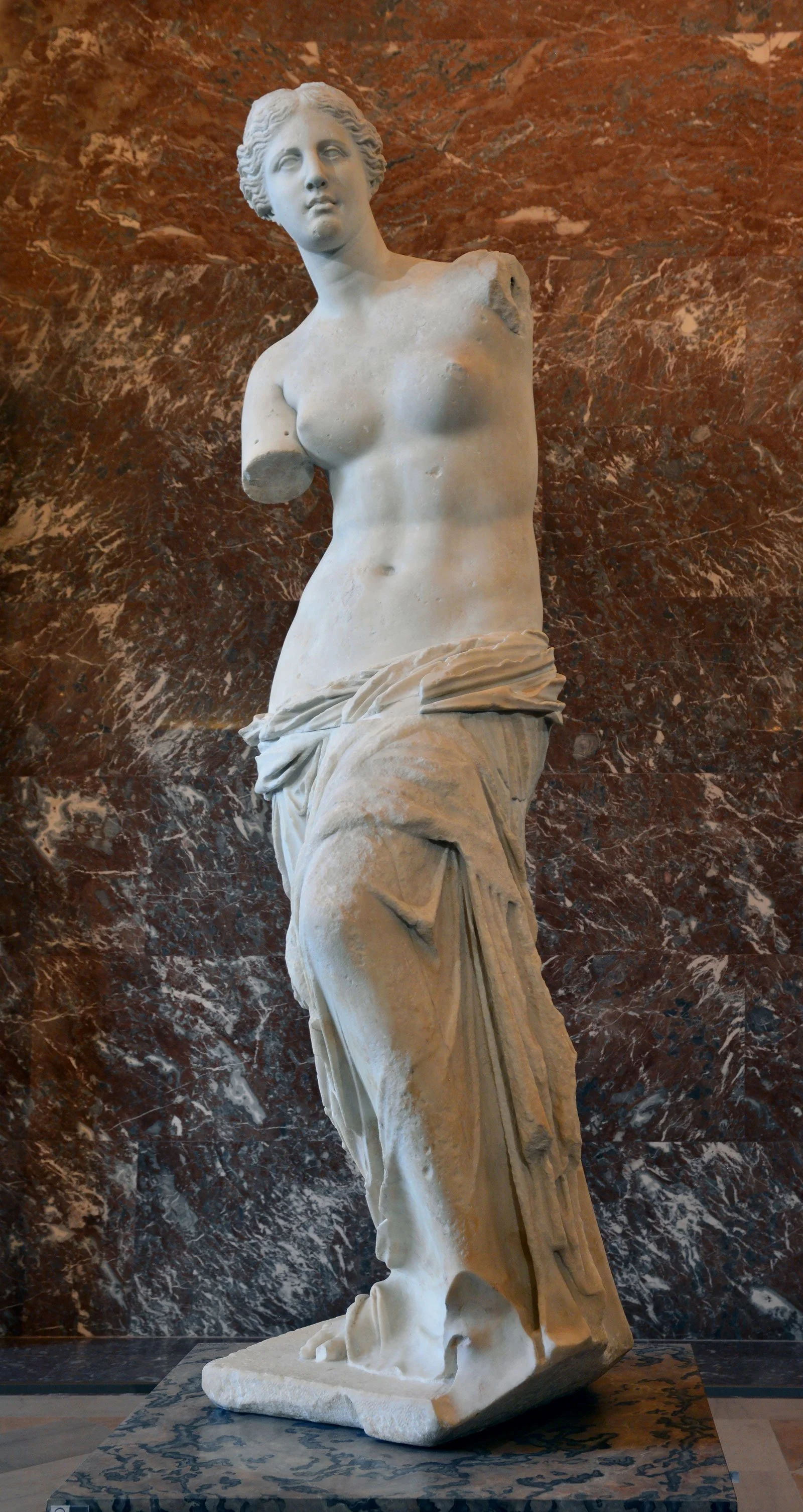 Front view of the Venus de Milo standing before a red marble wall.