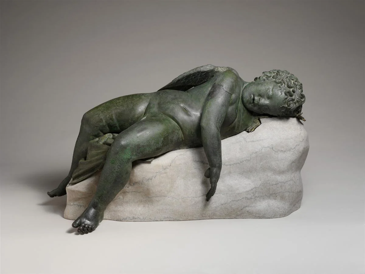 Bronze statue of a sleeping winged child lying on a stone base.
