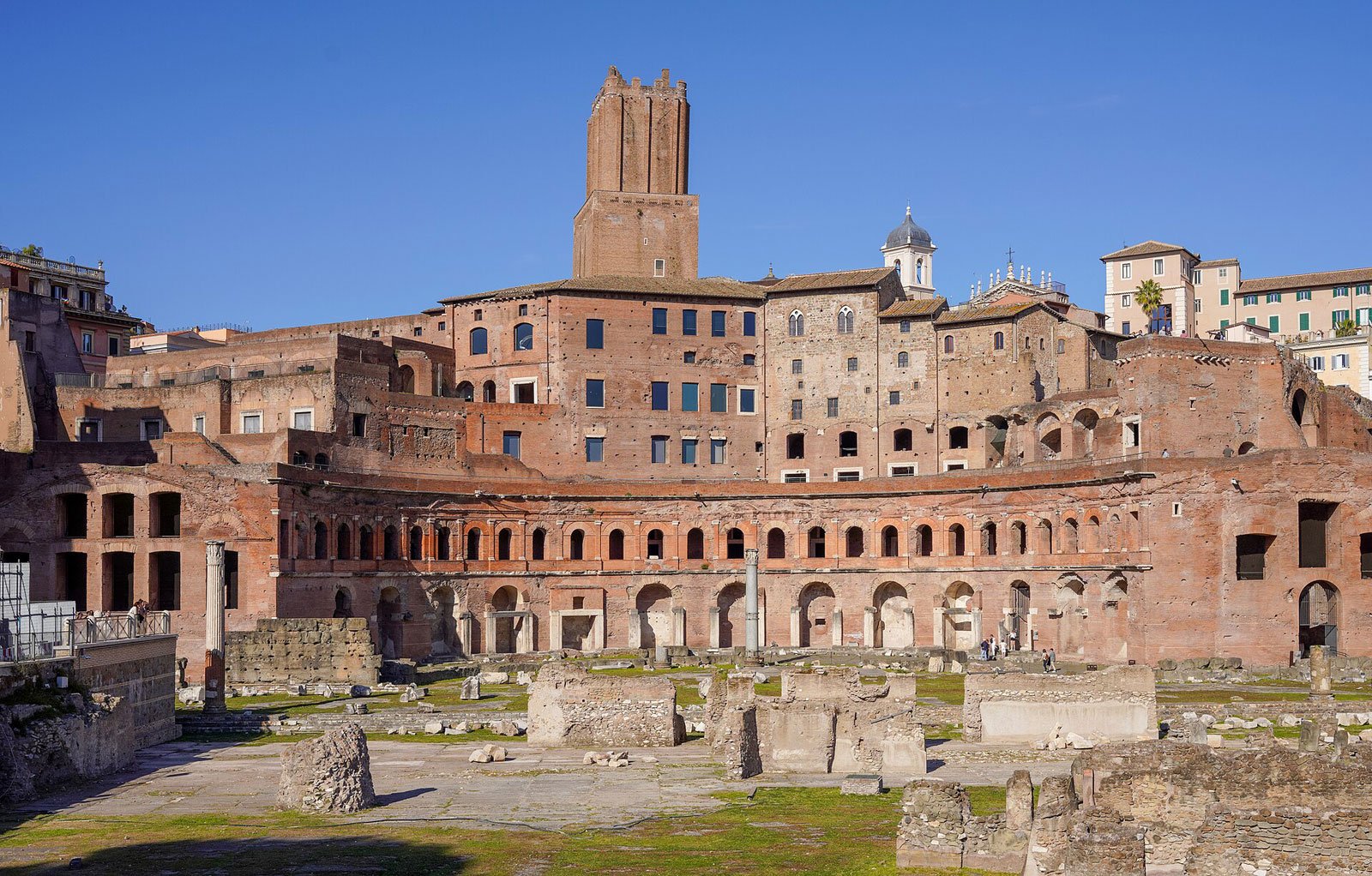 Exterior view of Trajan’s Market in Rome with layered brick arcades, ruins, and the tower rising above the complex.