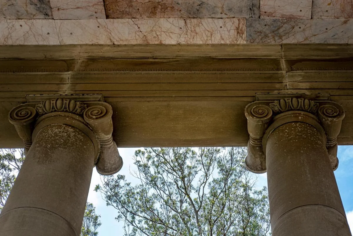Underside of a neoclassical portico, painted coffered ceiling and richly coloured Ionic capitals framing trees and bright sky.