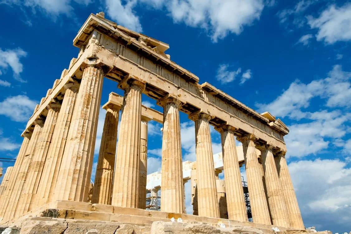 Wide view of the Parthenon’s Doric colonnade under a bright blue sky, weathered marble columns rising from the Acropolis rock.