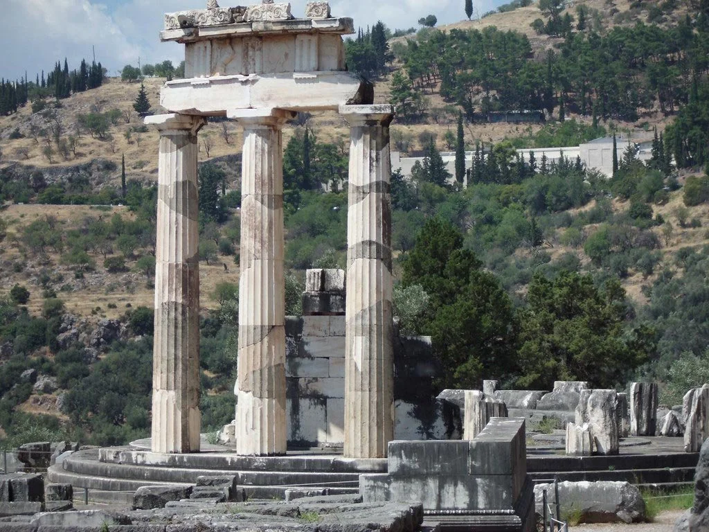 Circular tholos at Delphi with three restored Doric columns, standing amid scattered blocks against a wooded mountain slope.