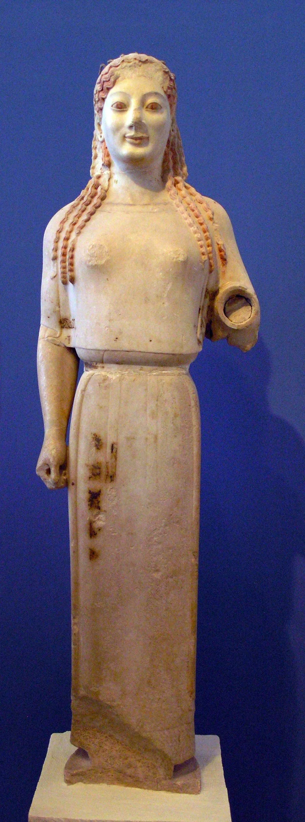 Original marble kore statue against a blue background, with stylised hair, archaic smile and faint surviving traces of surface colour.