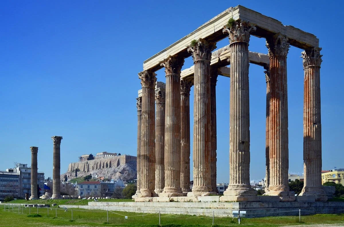 Ruined Temple of Olympian Zeus in Athens, tall Corinthian columns rising from a grassy field with the Acropolis in the distance.