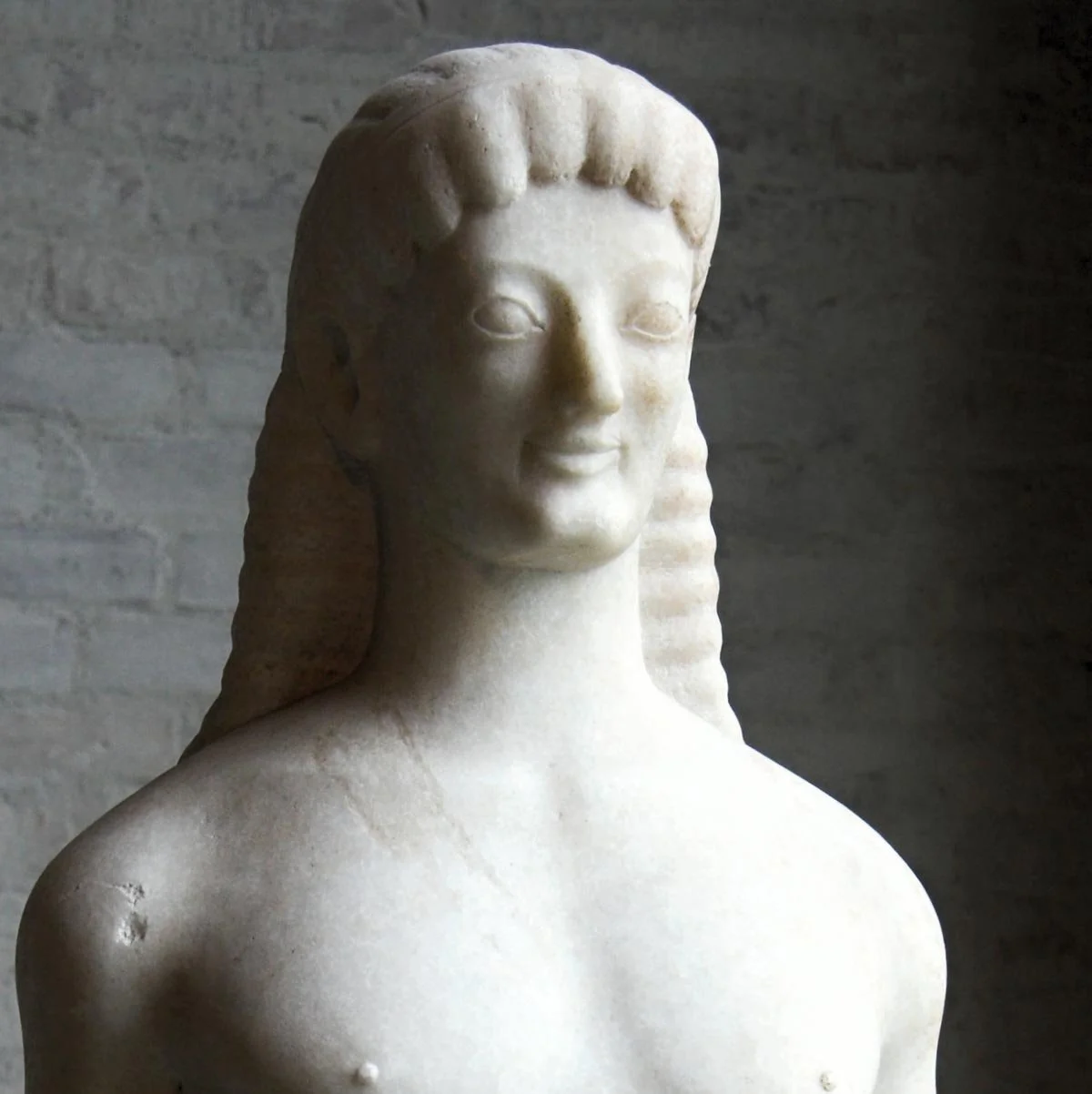 Archaic kouros statue with soft muscles and patterned hair, standing nude in the museum, one leg forward on a simple base.