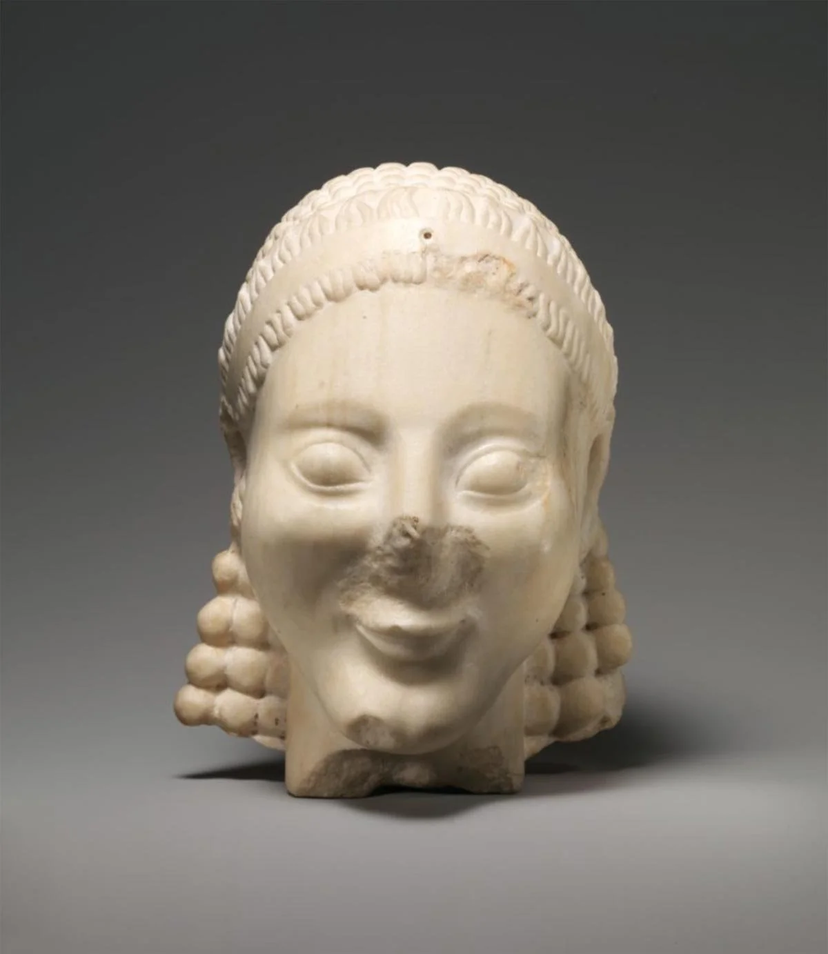Broken marble head of an archaic kouros, with stylized curls and archaic smile, nose damaged yet expression serene.