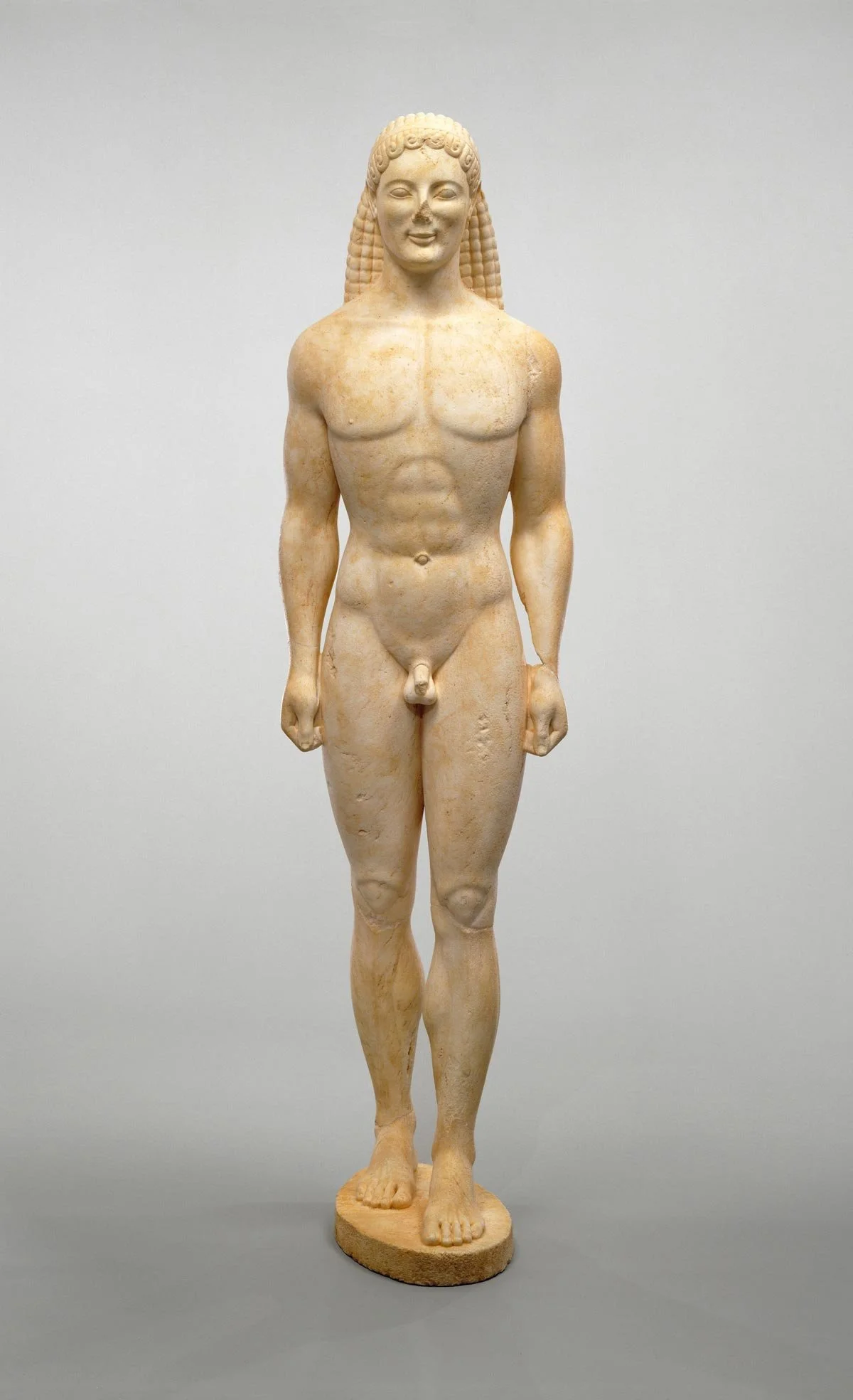 Full-length marble kouros stands frontally, one foot advanced, arms rigid at sides, body carved with idealized muscles.