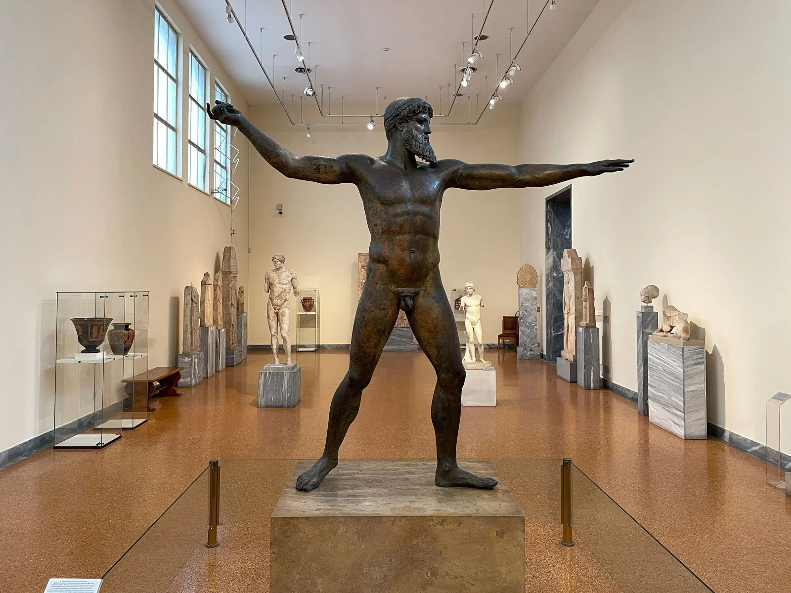 Artemision Bronze displayed in a museum gallery, arms extended across the room.