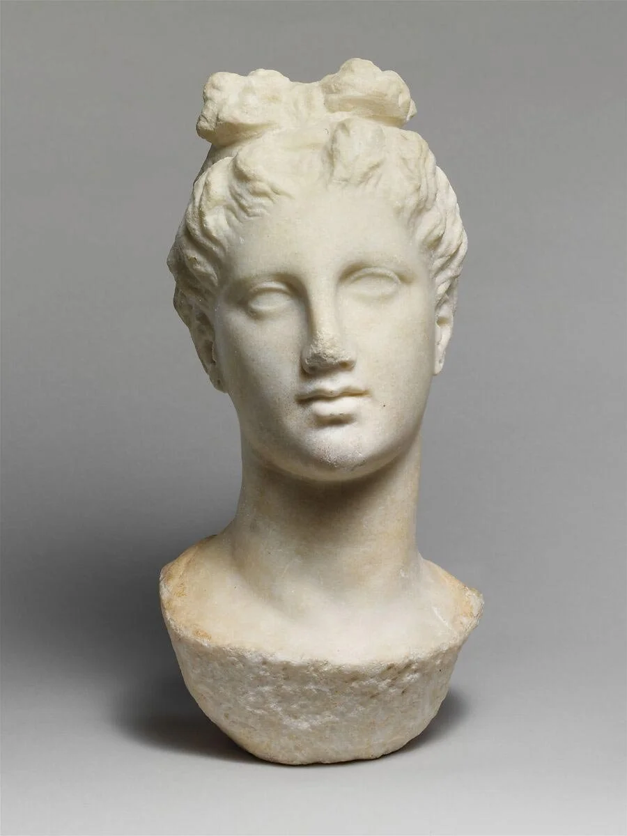 Marble head of a young woman with smooth features and hair gathered up from the face.