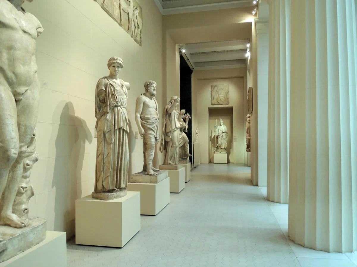 Cast gallery with rows of full-length classical statues in a high museum hall, lit from above and bordered by tall columns.